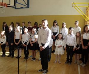 Uczeń klasy siódmej w stroju galowym recytuje przy mikrofonie. W tle pozostali uczniowie biorący udział w występie