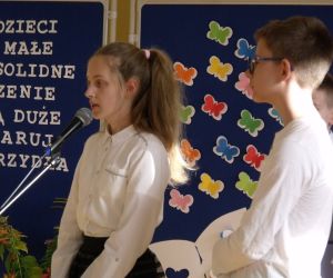 Uczennica i uczeń klasy siódmej w strojach galowych przy mikrofonie