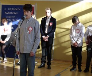 Uczeń klasy ósmej w stroju galowym przy mikrofonie. W tle pozostali występujący uczniowie i tablica dekoracyjna