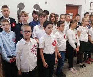 Uczniowie klasy czwartej i siódmej w strojach galowych i z kotylionami narodowymi śpiewają hymn państwowy