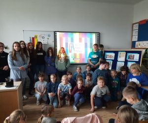 Uczniowie w niebieskich ubraniach stoją na tle prezentacji