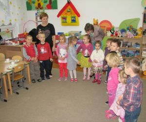 Dzieci z opiekunkami, stoją w klasie lekcyjnej. W tle widać materiały edukacyjne oraz zabawki.