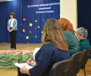 Zdjęcie przedstawia recytującą dziewczynkę na scenie i wsłuchaną komisję.