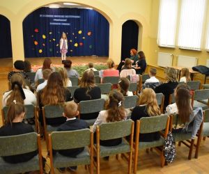 Zdjęcie przedstawia osoby siedzące na krzesłach, które słuchają występu dziewczynki stojącej na scenie.