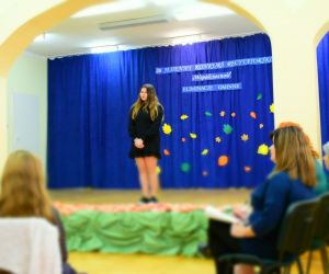 Zdjęcie przedstawia recytującą dziewczynkę na scenie i wsłuchaną komisję oceniającą.