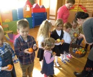 Dzieci stoją w rzędzie, nauczycielka częstuje je owocami