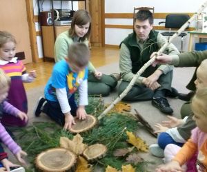 Dzieci siedzą w kole na podłodze razem z leśnikami i panią nauczycielką, jedno z nich trzyma plaster drewna