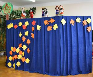 Większą część  zdjęcia zajmuje parawan koloru niebieskiego, do którego poprzyczepiane są żółte i pomarańczowe liście z papieru. Nad parawanem znajduje się sześć kukiełek: trzy Krakowianki, dwóch Krakowiaków oraz kukiełka Kata. Do dekoracji parawanu użyto drzewa wykonanego z papieru.