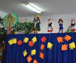 Na zdjęciu znajduje się parawan koloru niebieskiego, do którego poprzyczepiane są żółte i pomarańczowe liście z papieru. Nad parawanem znajdują się cztery kukiełki: dwie Krakowianki, jeden Krakowiak oraz Król. Do dekoracji parawanu użyto drzewa wykonanego z papieru. 