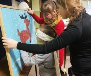 Na zdjęciu znajdują się trzy osoby oraz tablica, na której na niebieskim tle namalowany jest renifer. Dziewczynka przy pomocy animatora przyczepia ogonek do renifera.