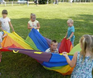 Grupa dzieci bawi się przy użyciu chusty animacyjnej, która jest różnokolorowa. Dzieci trzymają chustę za specjalne uchwyty, a w środku w chuście siedzi chłopczyk, który się uśmiecha