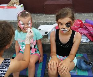 Na zdjęciu animator maluje twarze 2 dziewczynkom. Jedna ma namalowanego pieska, a druga motylka.