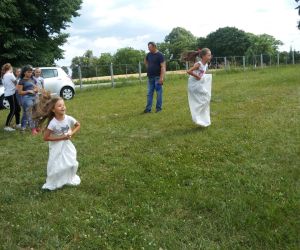 Na zdjęciu dwie dziewczynki skaczą w workach, podczas konkurencji sportowej. W oddali grupka dzieci kibicuje dziewczynkom.