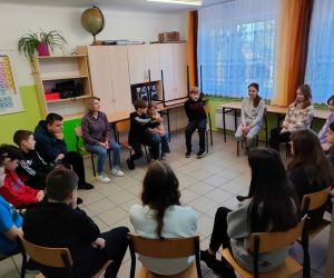 Grupa osób siedzi w kole wewnątrz budynku.