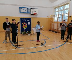 Chłopcy stoją przed mikrofonami, w tle dekoracja z okazji Dnia Kobiet