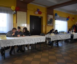 Zdjęcie przedstawia dzieci siedzące przy stołach wewnątrz budynku.