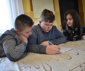 Zdjęcie przedstawia troje dzieci, jedno z nich pisze na kartce.