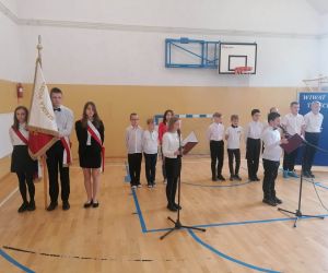 Poczet sztandarowy i dwoje uczniów klasy IV przy mikrofonach. W tle pozostali uczniowie klasy IV