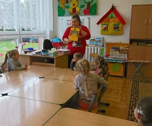 Zdjęcie przedstawia dzieci siedzące w ławkach słuchające czytanej przez panią książki.