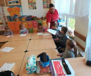 Zdjęcie przedstawia dzieci siedzące w szkolnych ławkach i panią która rozdaj im kolorowanki.