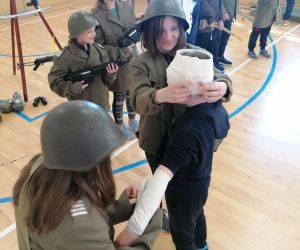 Dwie uczennice w umundurowaniu bandażują rękę ucznia przebranego za żołnierza