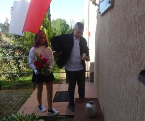 Uczeń i uczennica przy tablicy patrona szkoły składają kwiaty i zapalają znicz