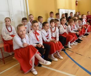 Grupa uczniów w ludowych strojach siedzi na ławce przed występem