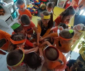 Dzieci stoją w kole przy stoliku, na którym znajduje się dynia. Wspólnie wyjmują ziarna dyni i oddzielają je od miąższu.