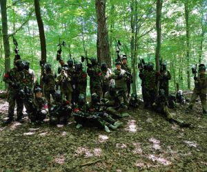 Uczniowie i nauczyciele w mundurach polowych, trzymają w dłoniach karabinki do gry w paintball. Scena w lesie