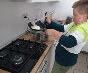 Chłopiec w biało-zielonej bluzie wlewa ciasto naleśnikowe na patelnię