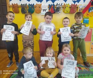 Dzieci prezentują się z dyplomami z okazji Dnia Kota. W tle prace plastyczne przedstawiające koty.