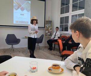 Kobieta w kapeluszu recytuje na tle ekranu z prezentacją