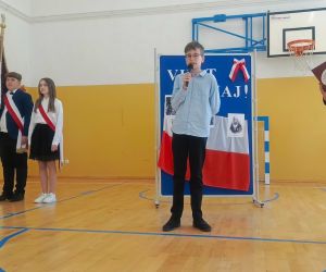 Uczeń klasy czwartej i uczeń klasy siódmej przy mikrofonach, w tle poczet sztandarowy