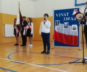 Uczeń klasy czwartej i uczeń klasy siódmej przy mikrofonach, w tle poczet sztandarowy