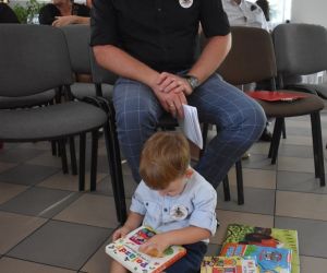 Na zdjęciu widoczne jest czytające książki dziecko i mężczyzna siedzący na krześle.