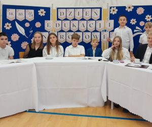 Uczniowie klasy szóstej przy stole na tle dekoracji okolicznościowej