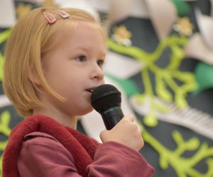 Dziewczynka na scenie wewnątrz budynku, śpiewa do mikrofonu.