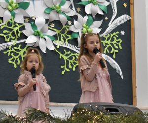 Dziewczynki na scenie wewnątrz budynku, śpiewają do mikrofonu.