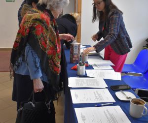 Osoby wewnątrz budynku, przy stoliku wypełniają dokumenty.